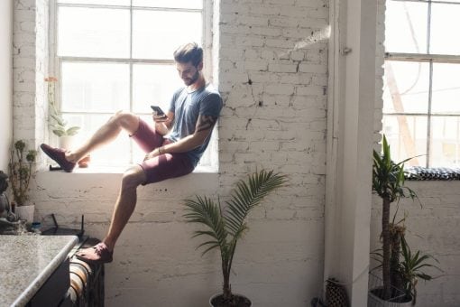 Young man sits on a windowsill in his apartment looking down at his mobile phone.
