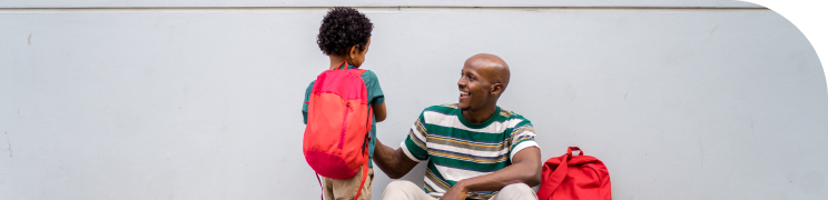 A man sits on a bench as he smiles at a young boy with a red backpack standing next to him.