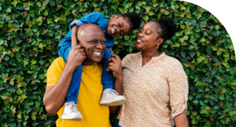 Smiling family with a young child on the father's shoulders, standing in front of a lush green ivy-covered wall