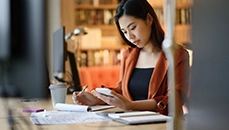 Woman seated at desk looks at a calculator looks at a calculator in her left hand.